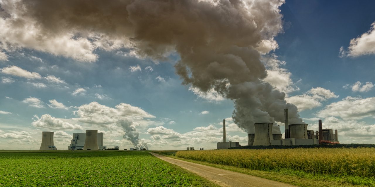 Industrial power plant emitting thick smoke into the sky, situated next to green farmland, symbolizing the contrast between industrial pollution and agriculture | Landmark Judgment on Climate Change, Carbon Markets, Offset Projects, Public Participation, and Community Land Rights in Kenya | ESG and climate change lawyer in Nairobi | Njaga & Co. Advocates LLP ESG law firm