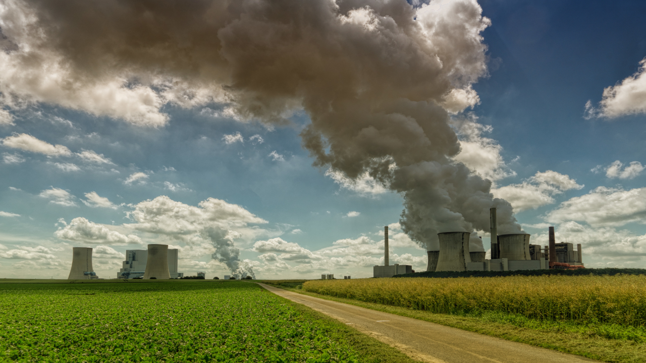 Industrial power plant emitting thick smoke into the sky, situated next to green farmland, symbolizing the contrast between industrial pollution and agriculture | Landmark Judgment on Climate Change, Carbon Markets, Offset Projects, Public Participation, and Community Land Rights in Kenya | ESG and climate change lawyer in Nairobi | Njaga & Co. Advocates LLP ESG law firm